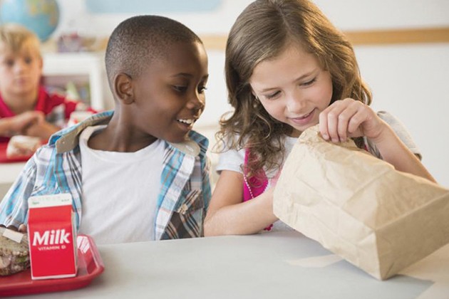 Two children peeking into a brown bag at the school lunch table.