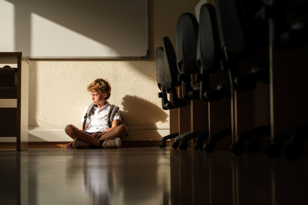 Little boy sitting sadly against a wall in a classroom.